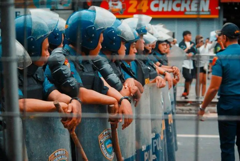 A line of police officers in riot gear standing on a street during a public gathering.